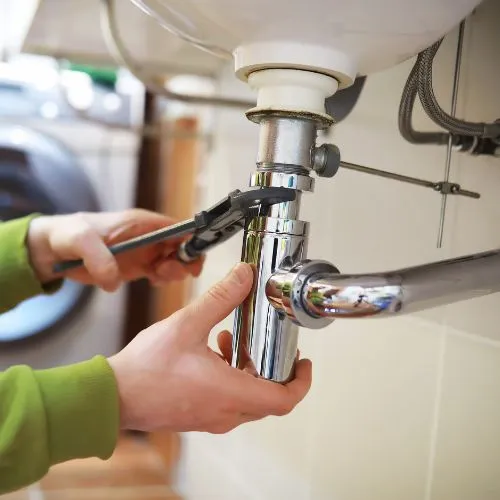 Un plombier resserre un siphon chromé sous un lavabo avec une clé, lors d'un dépannage de fuite
